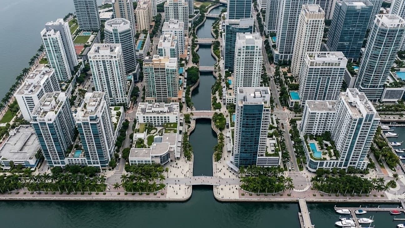 A wide aerial editorial photograph of a mixed-use waterfront development with distinctive high-rise towers and canals at overcast midday, small distant pedestrians crossing a plaza far below, no legible text or logos.