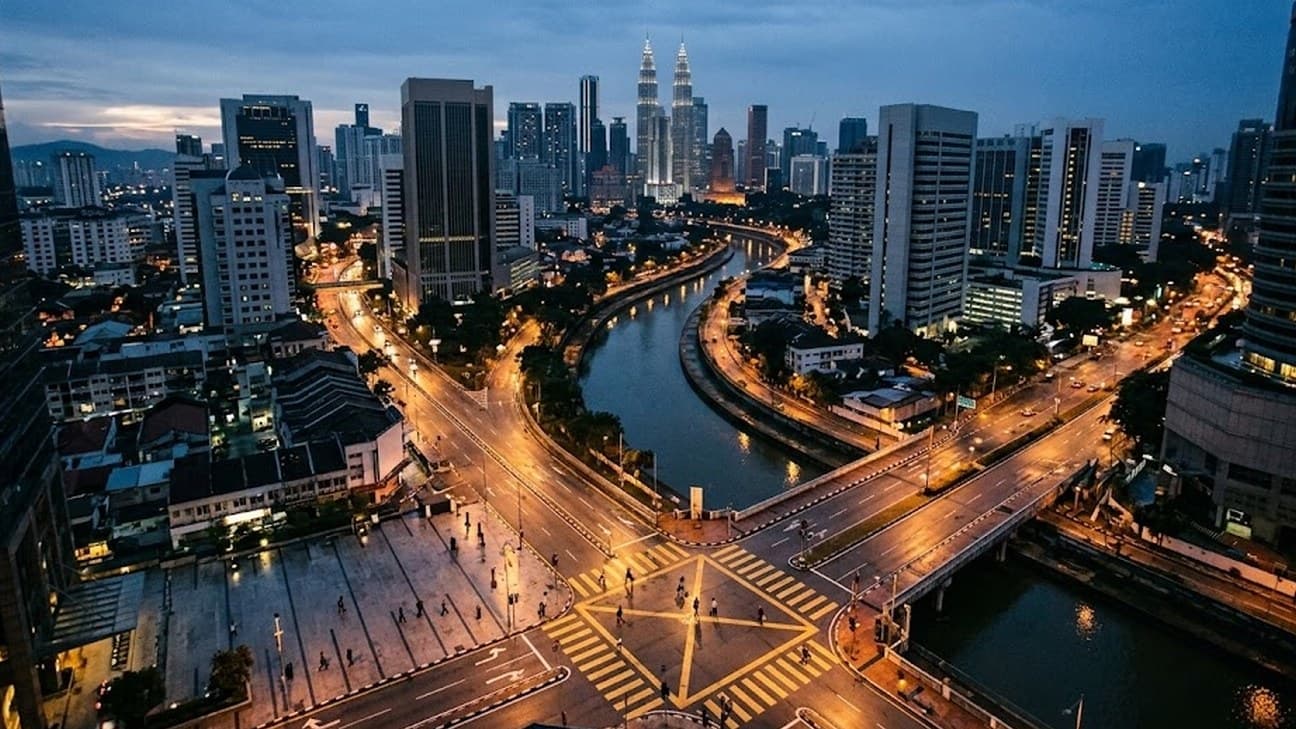 High-angle view of a Kuala Lumpur financial district at pre-dawn blue hour, with office towers, a river bend and a few tiny pedestrians crossing a floodlit plaza and a marked crosswalk.